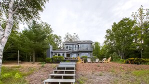 A two-story house with a balcony, surrounded by trees and a wooden staircase leading to the front door. - Lavish Charlevoix Home on Lake Michigan, Fire Pit (Charlevoix)