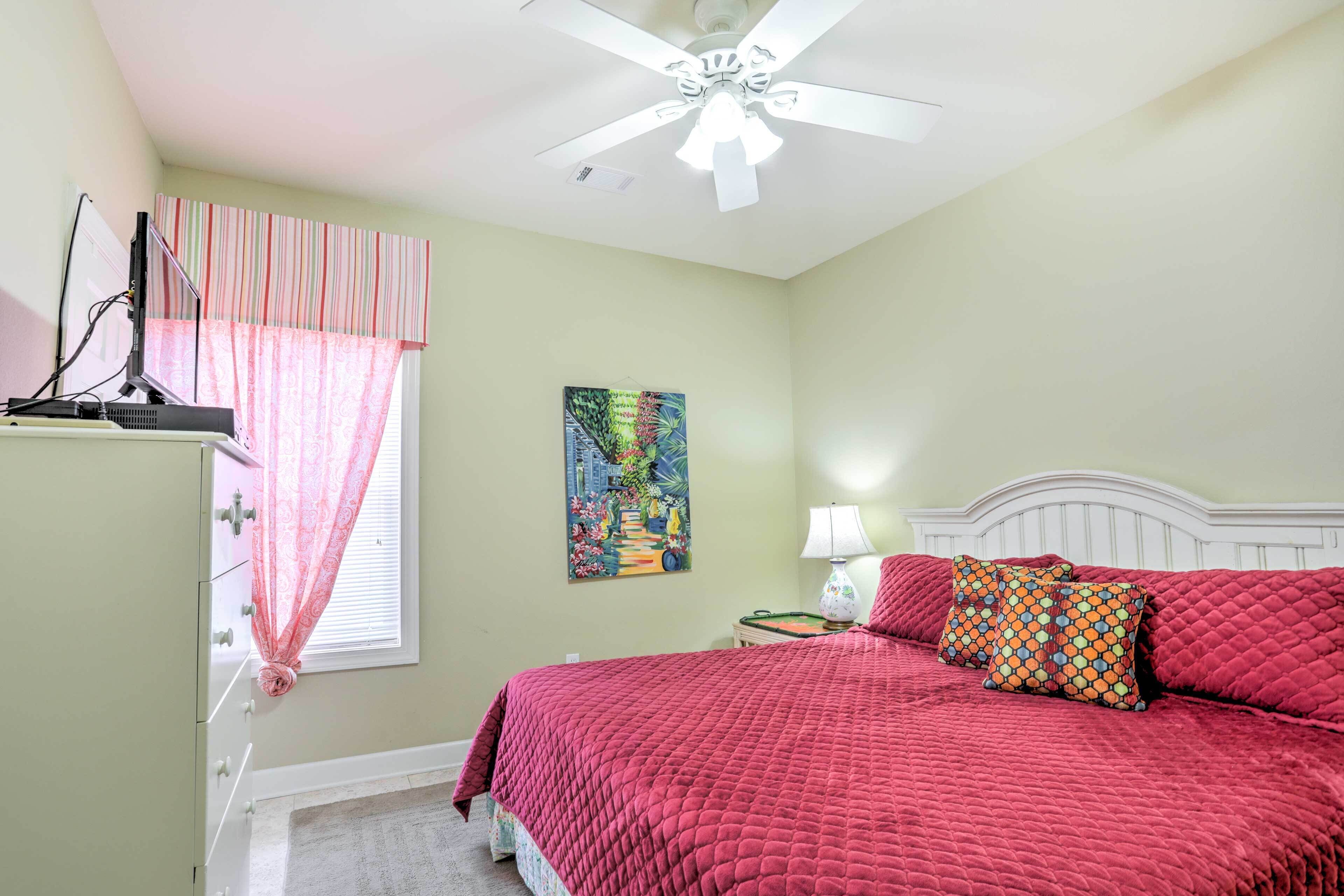 A bedroom with a bed, a ceiling fan, a window with curtains, a painting on the wall, and a nightstand with a lamp.