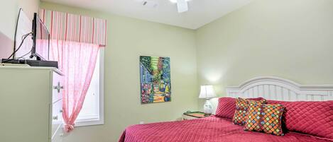 A bedroom with a bed, a ceiling fan, a window with curtains, a painting on the wall, and a nightstand with a lamp.