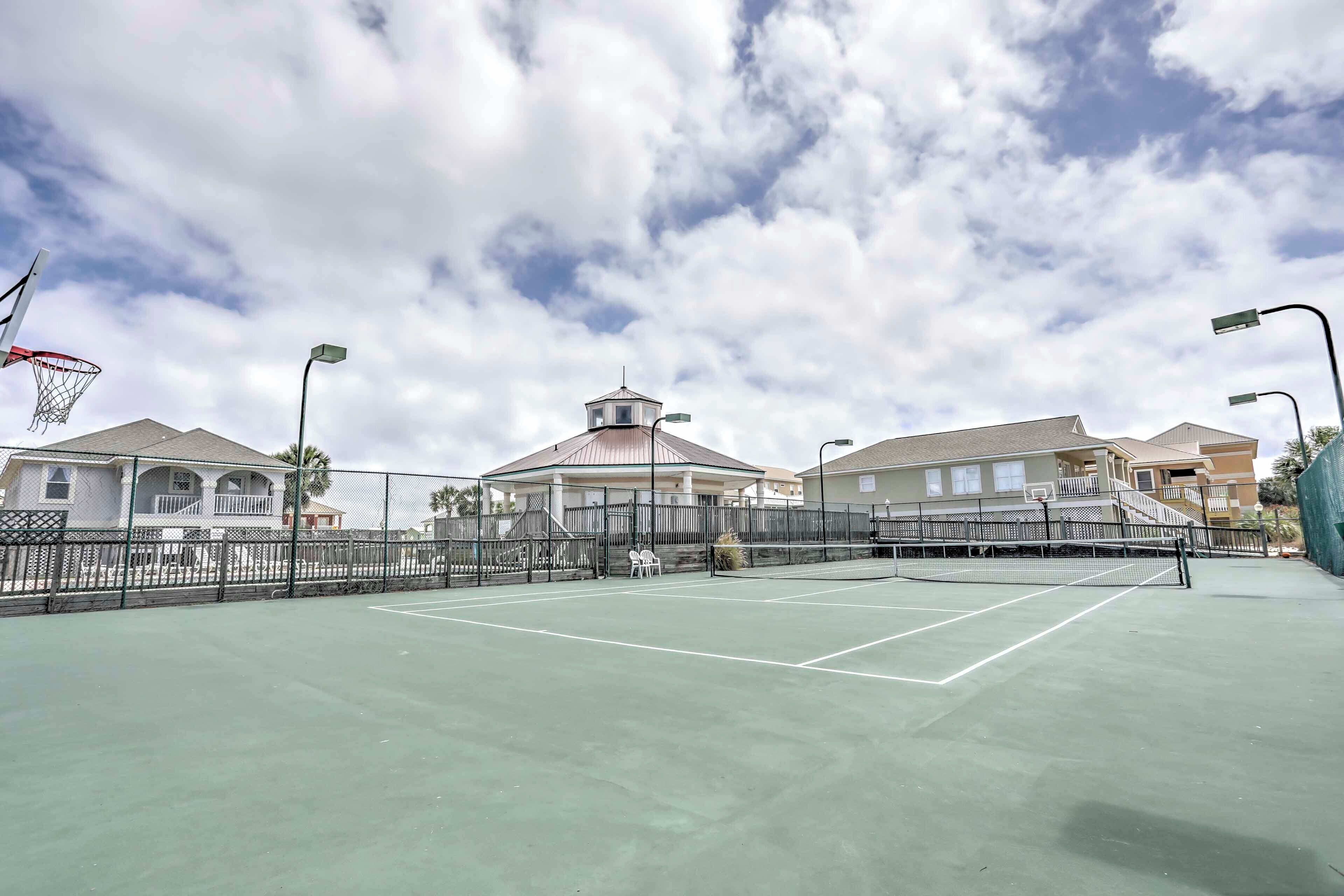 An outdoor tennis court with a net, surrounded by a fence, and adjacent to residential buildings.