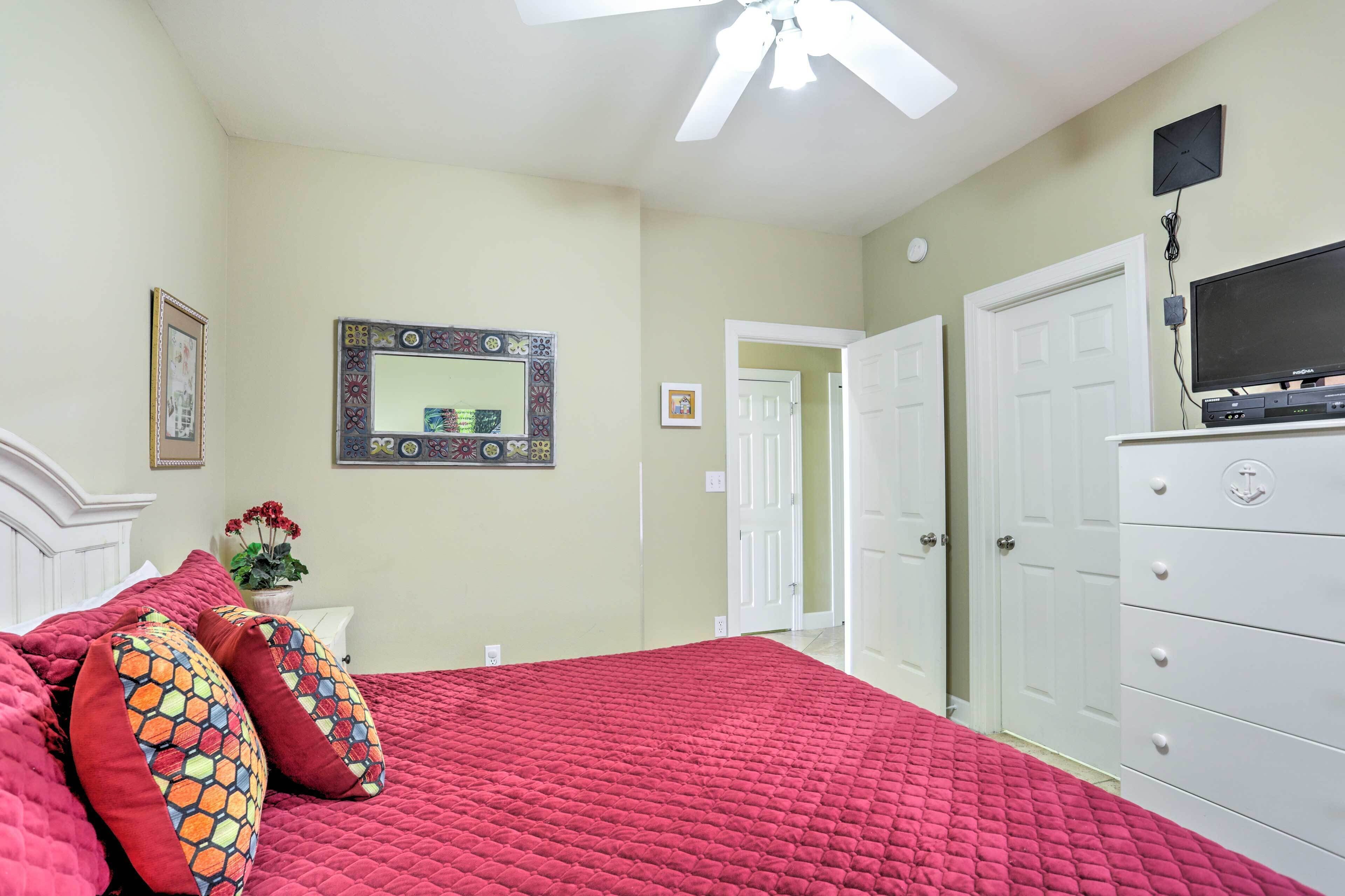 A bedroom with a red quilted bedspread, a white dresser, a ceiling fan, and a television on a stand.