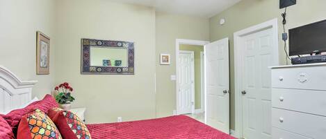 A bedroom with a red quilted bedspread, a white dresser, a ceiling fan, and a television on a stand.