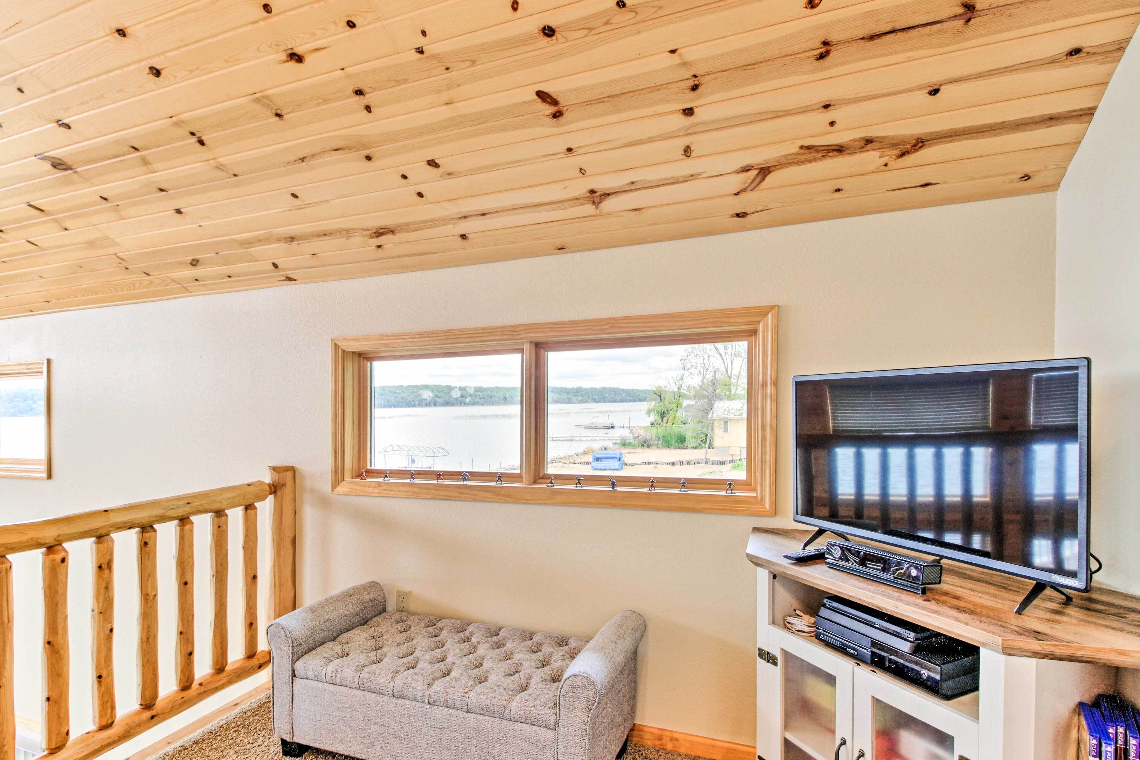 Una habitación con techo de madera, una ventana con vista al agua, un televisor sobre un soporte de madera y un banco acolchado.