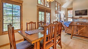 A dining area with a wooden table and chairs, large windows with blinds, a stone fireplace, and a television mounted on the wall.