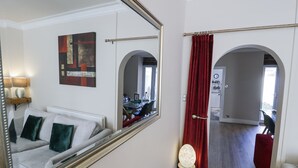 A hotel room with a large mirror, a red curtain, a sofa with green cushions, a small table with a lamp, and a view of the kitchen and dining area through an archway.