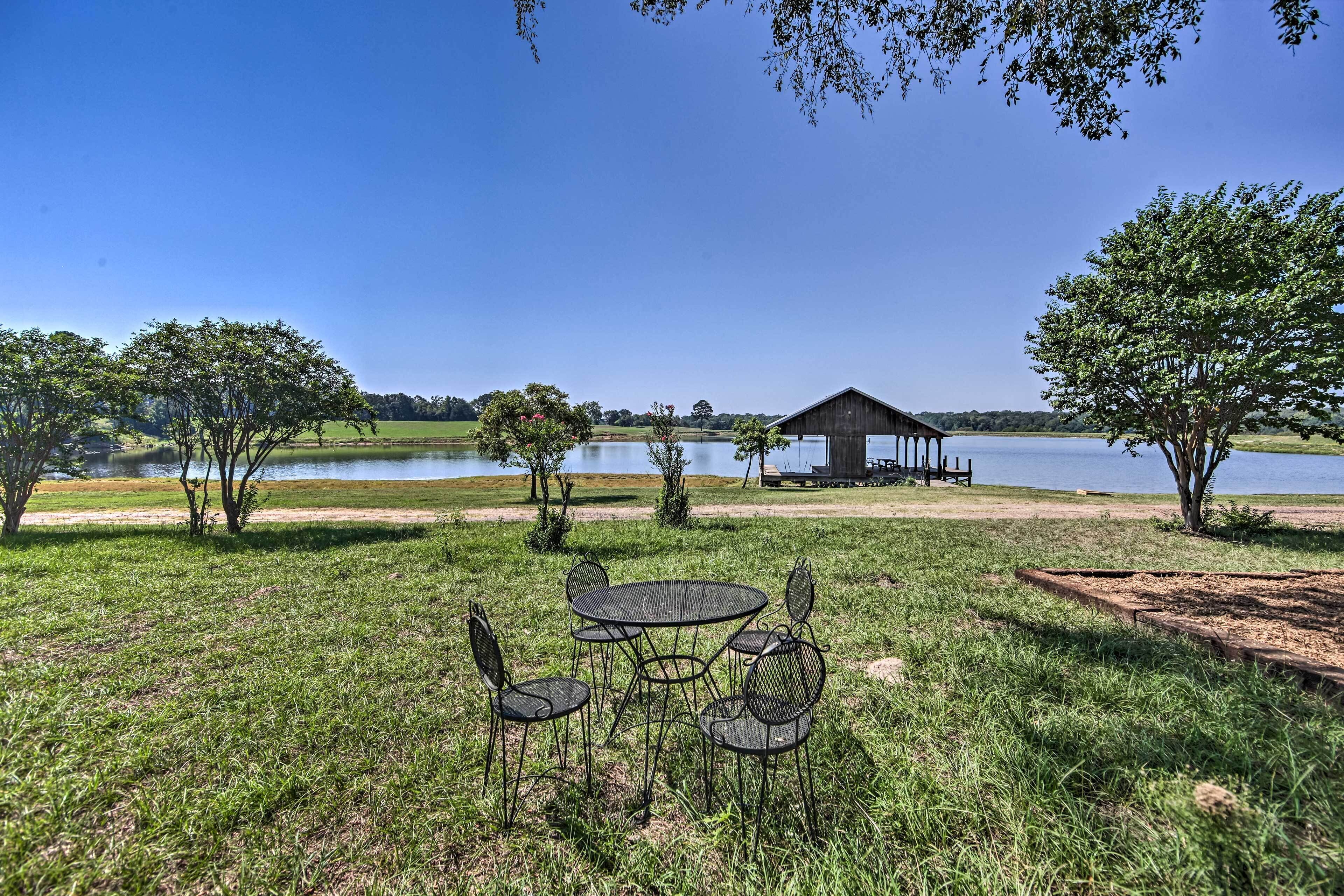 A grassy area with a metal table and chairs, a wooden structure by a lake, and trees around.