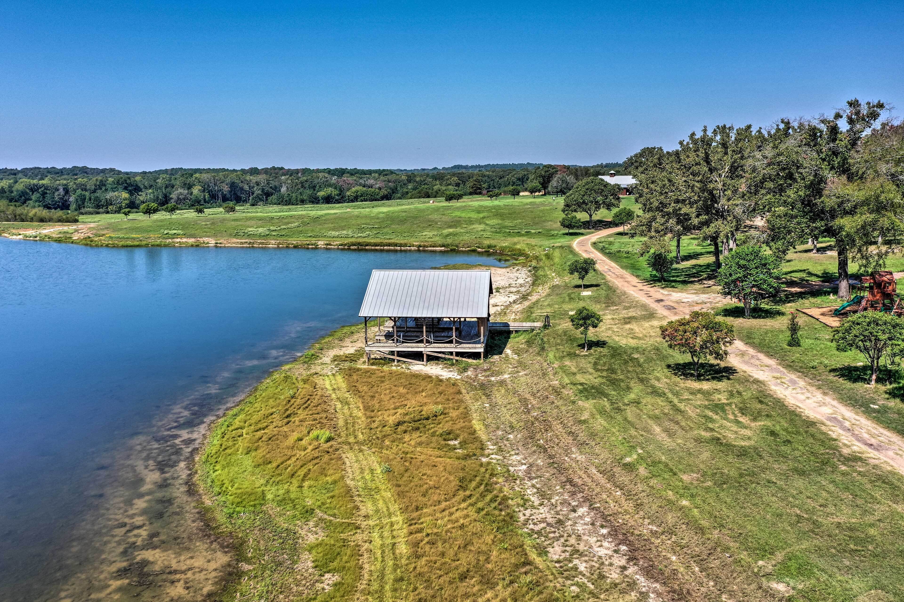 Una cabaña junto al lago con techo de metal, rodeada de campos verdes y árboles, cerca de un camino de tierra.