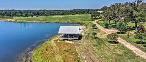 A lakeside cabin with a metal roof, surrounded by grassy fields and trees, near a dirt path.