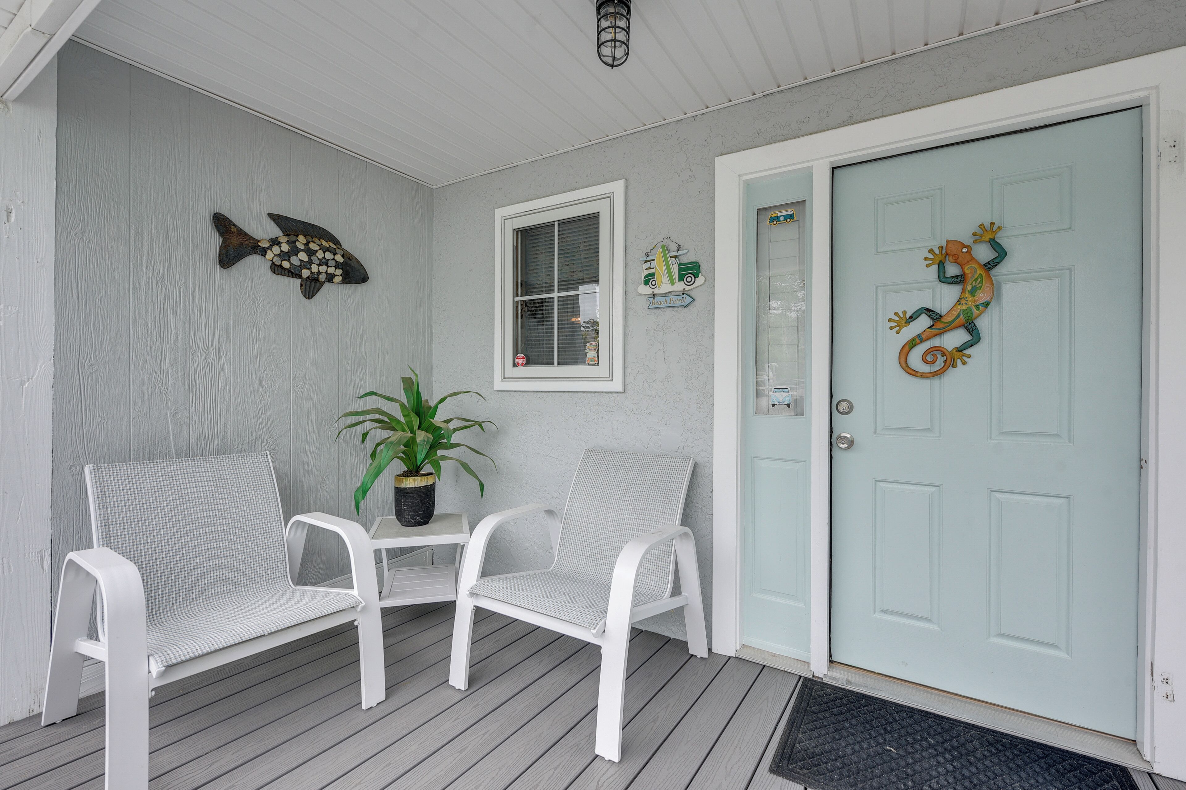 A porch with two white wicker chairs, a potted plant, a fish wall decoration, a window, and a door with a lizard decoration.
