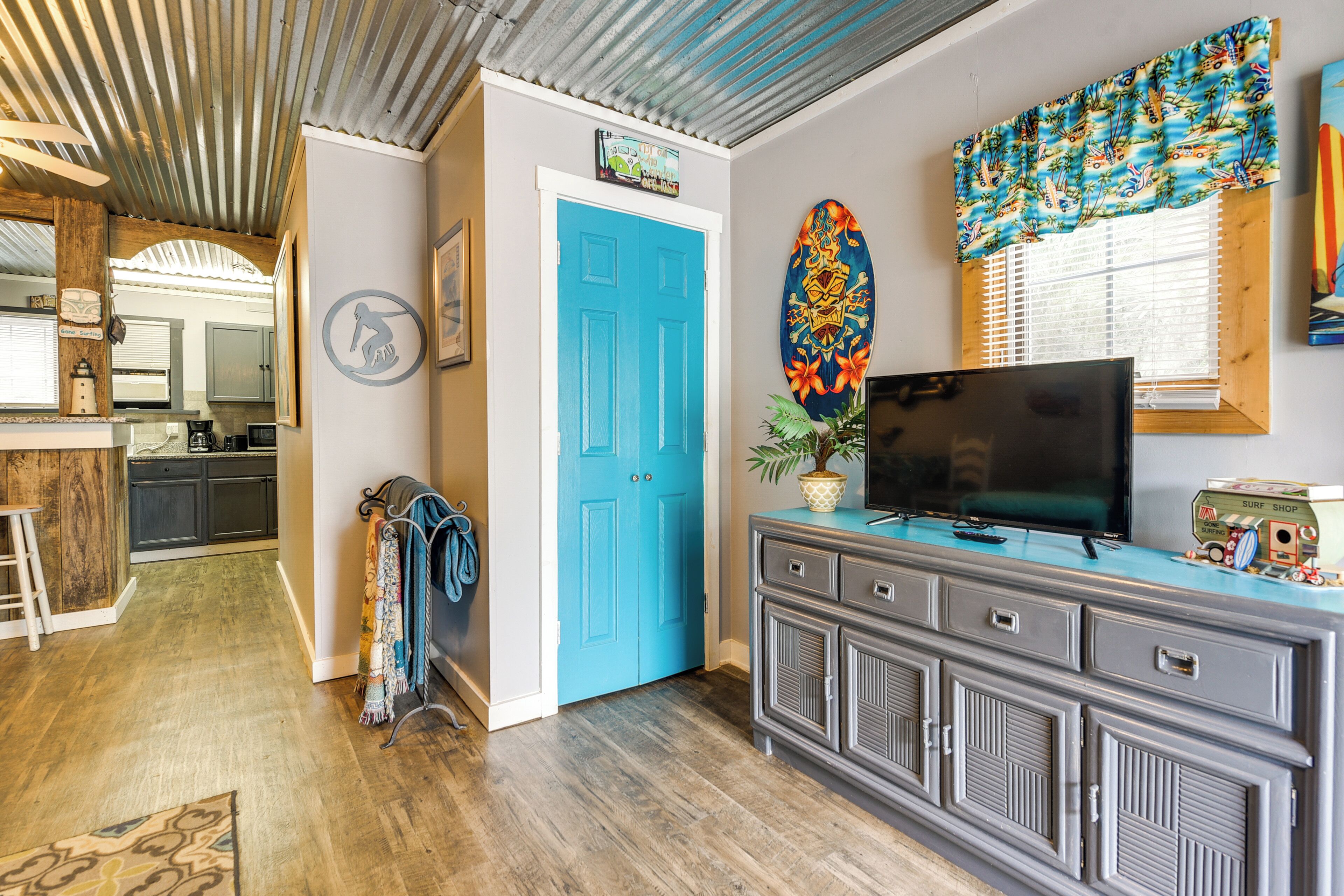 Una habitación con puerta azul, televisor sobre una cómoda, decoración con una tabla de surf y una cocina al fondo.