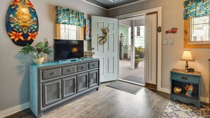 A room with a television on a cabinet, a door with a lizard decoration, a lamp on a side table, and a colorful artwork on the wall. - 1 Mi to Ocean Isle Beach! Studio w/ Outdoor Oasis (Ocean Isle Beach)