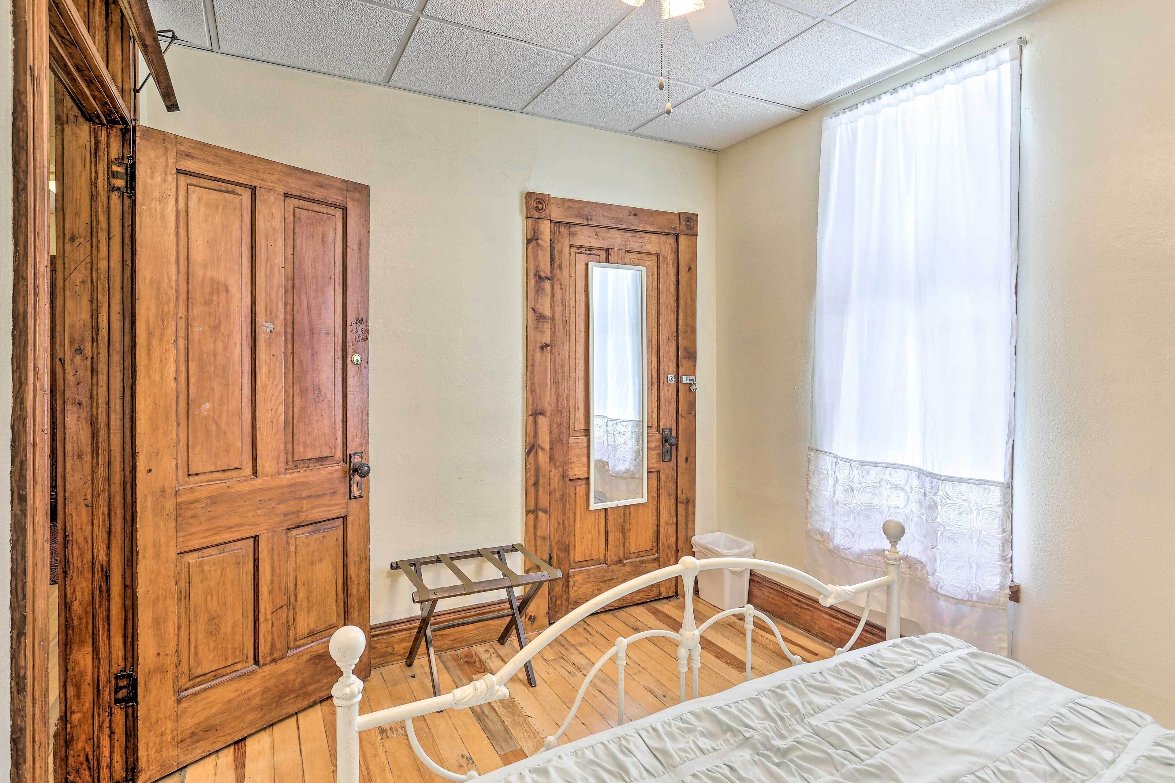 Une chambre avec une porte en bois, une petite fenêtre ornée de rideaux légers, un lit blanc et un plancher en bois.