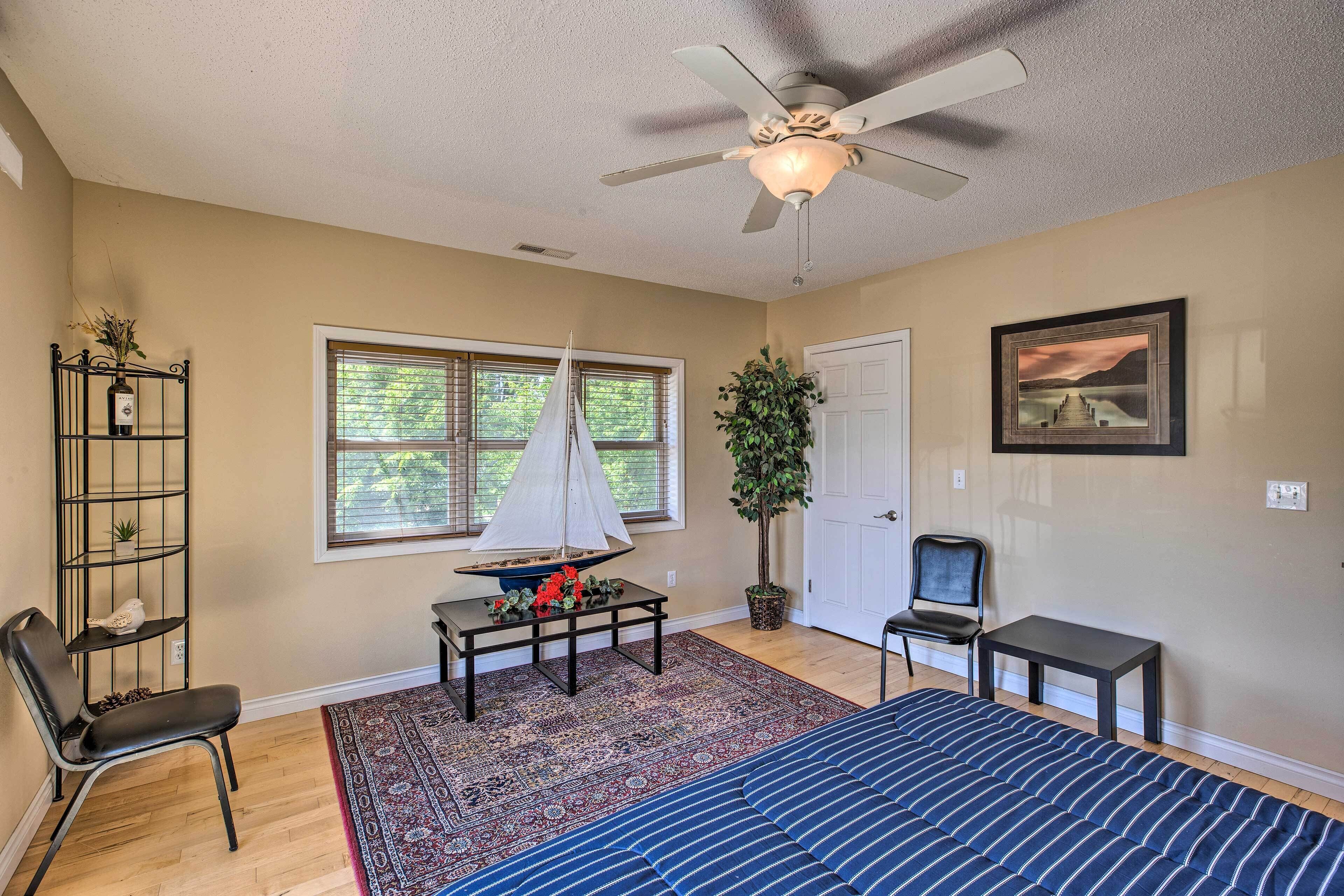 A bedroom with a bed, a ceiling fan, a window with blinds, a potted plant, a small table with decorative items, and a chair.
