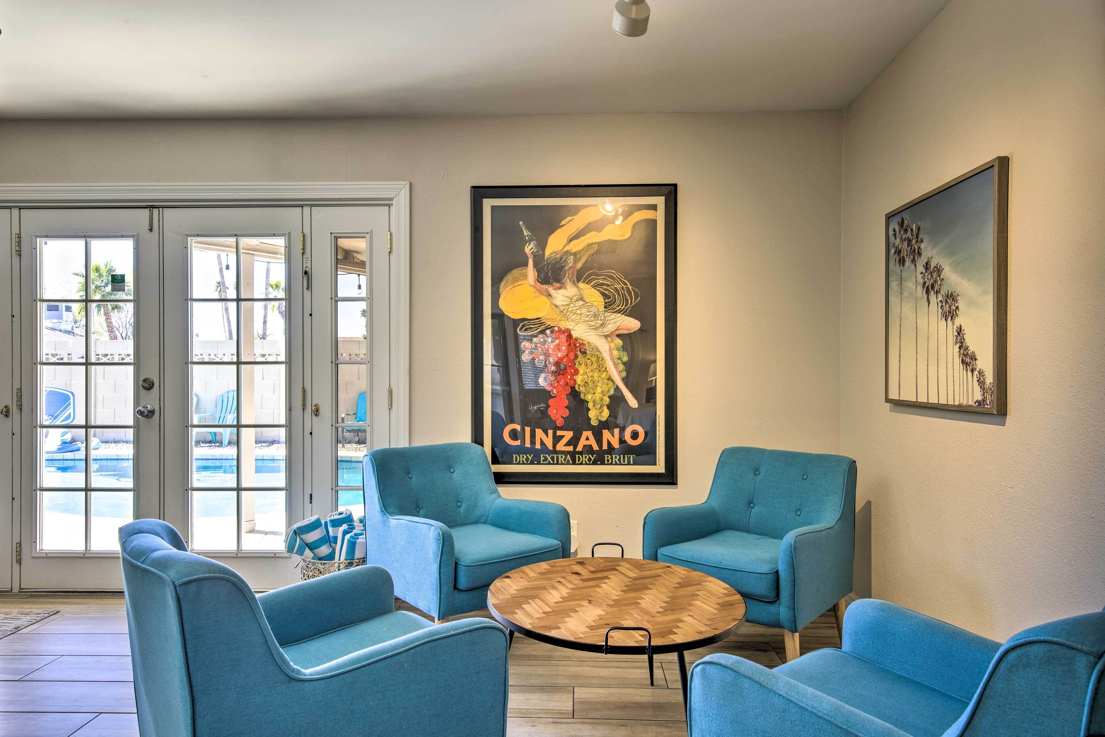 A hotel lobby with a large poster of a woman holding a bottle, two blue armchairs, a round wooden table, and a door leading outside.