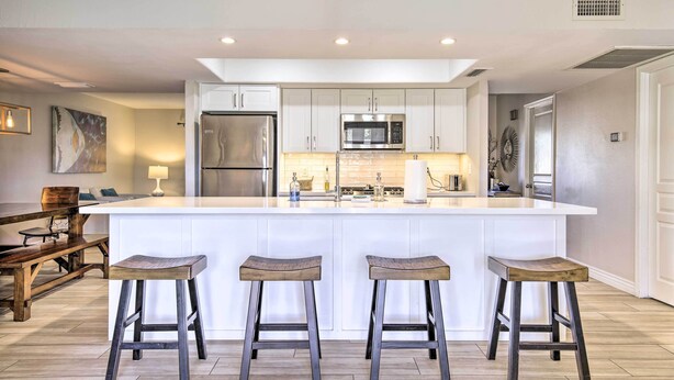 A modern kitchen with white cabinetry, a central island with stools, stainless steel appliances, and a wooden dining area. - 3 Mi to Asu: Tempe Getaway w/ Heated Pool (Tempe)