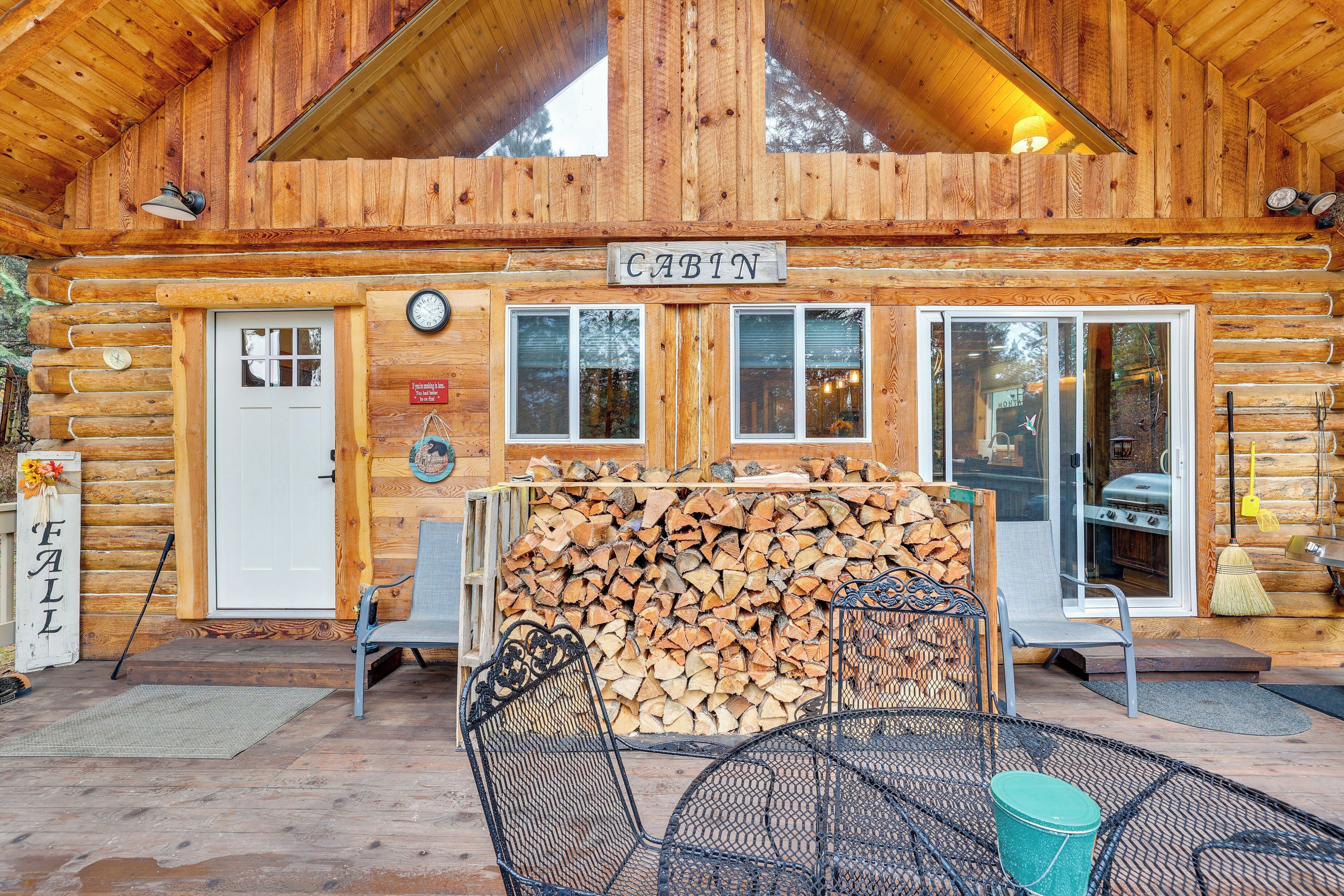 Une cabane en rondins avec une pancarte indiquant « Cabane », une porte, des fenêtres, une pile de bois de chauffage, des chaises en métal et une table.