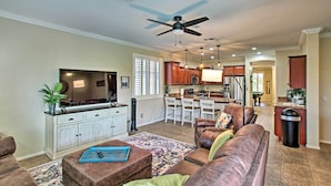 A spacious living room with a brown leather sofa, a coffee table, and a TV on a white cabinet. There is a kitchen area with wooden cabinets and a dining table. - Game Room, Fenced Yard: Estrella Oasis in Goodyear (Goodyear)