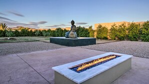 Una estructura de fuego en un macetero de piedra rectangular, una estatua sobre un pedestal y un jardín ajardinado con árboles y arbustos.