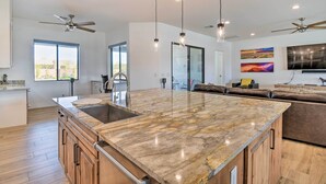 Una cocina moderna con una isla grande, armarios de madera y un lavamanos empotrado. Hay ventiladores de techo, lámparas colgantes y un ventanal que proporciona luz natural.