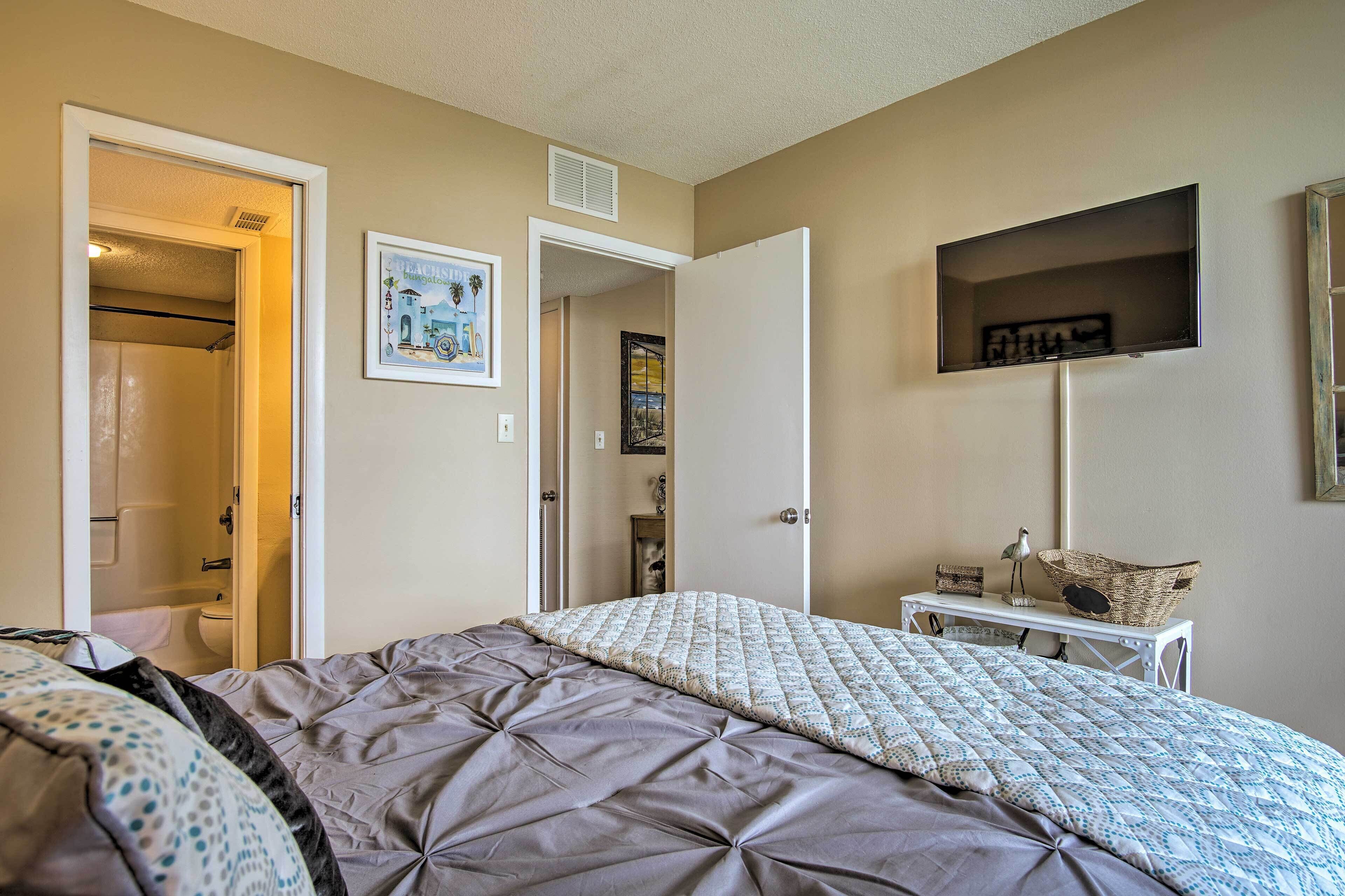 A bedroom with a bed, a bedside table with a basket, a wall-mounted TV, and a door leading to a bathroom.