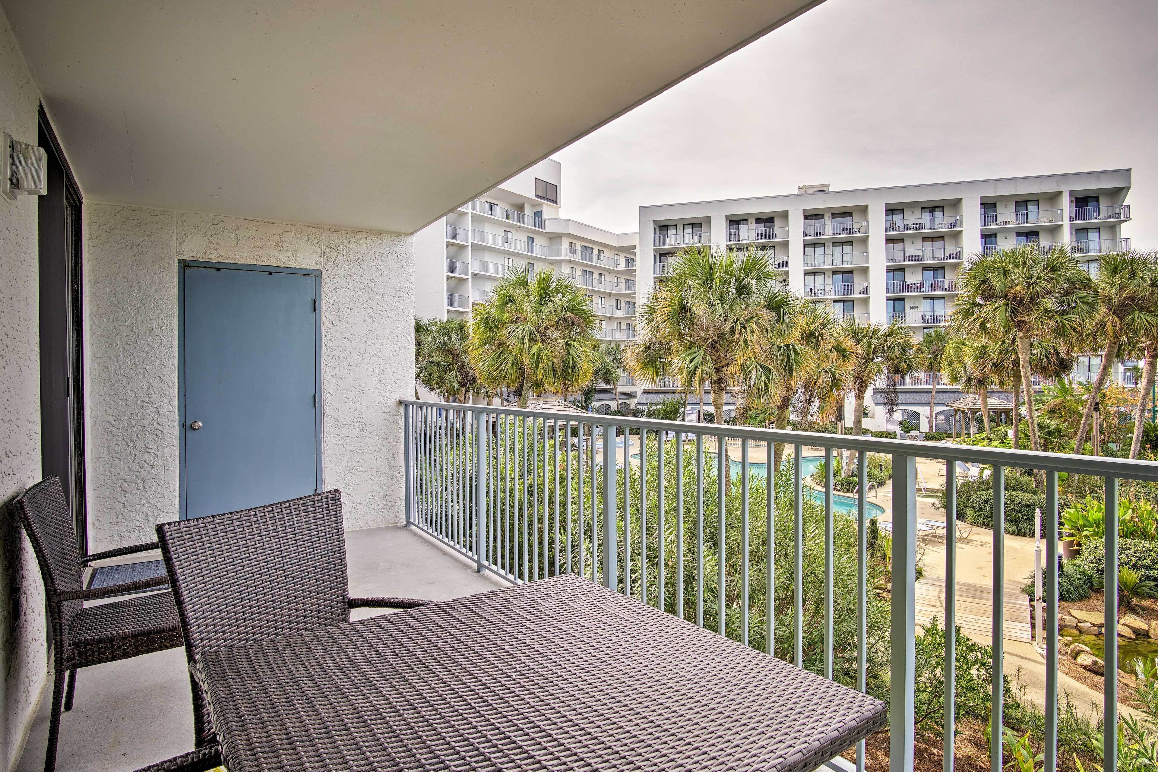 A balcony with wicker furniture, a blue door, and a view of a swimming pool and palm trees.