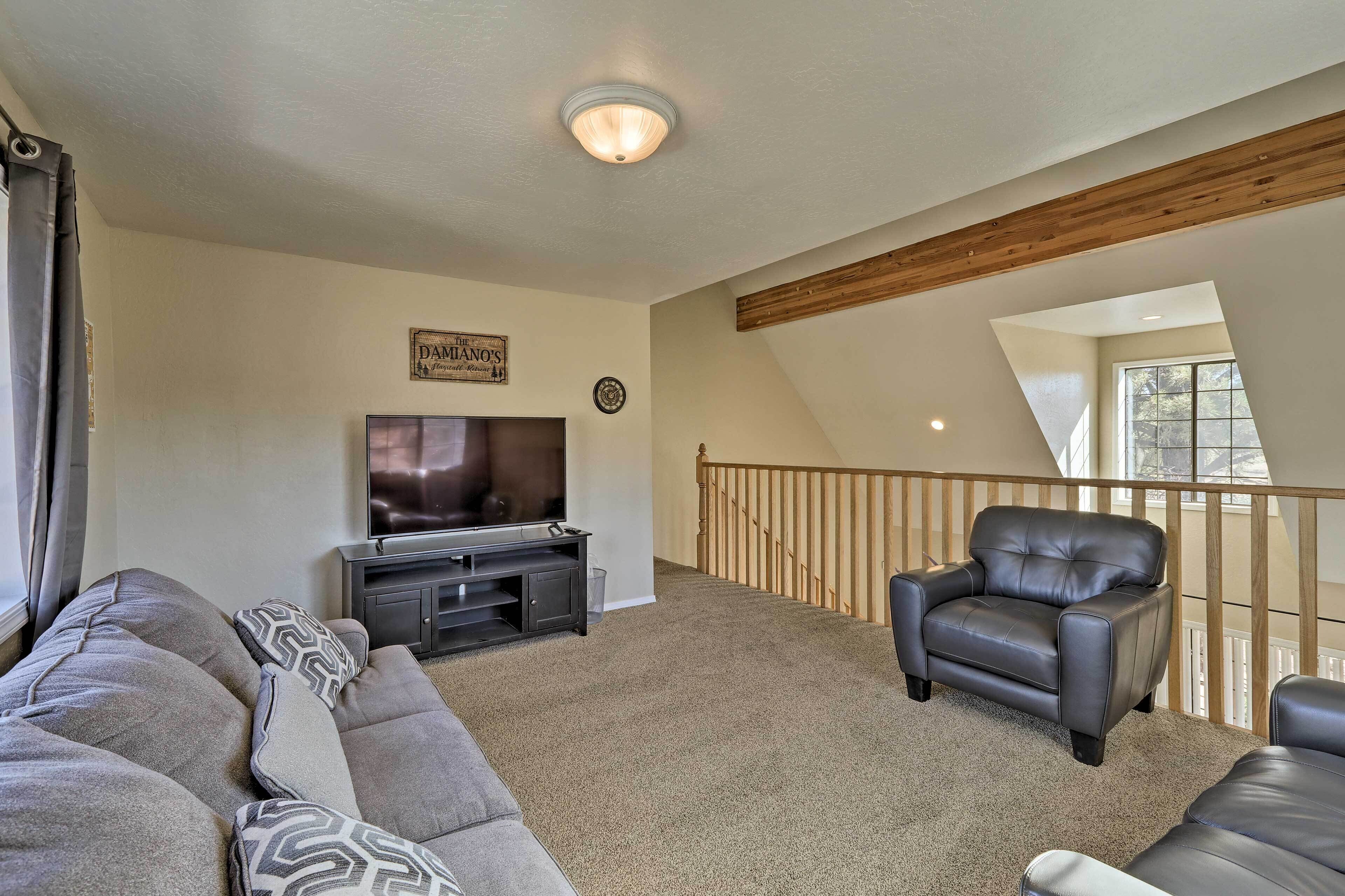 A living room with a large gray sectional sofa, a black leather armchair, a flat-screen TV on a black entertainment unit, and a wooden ceiling beam.