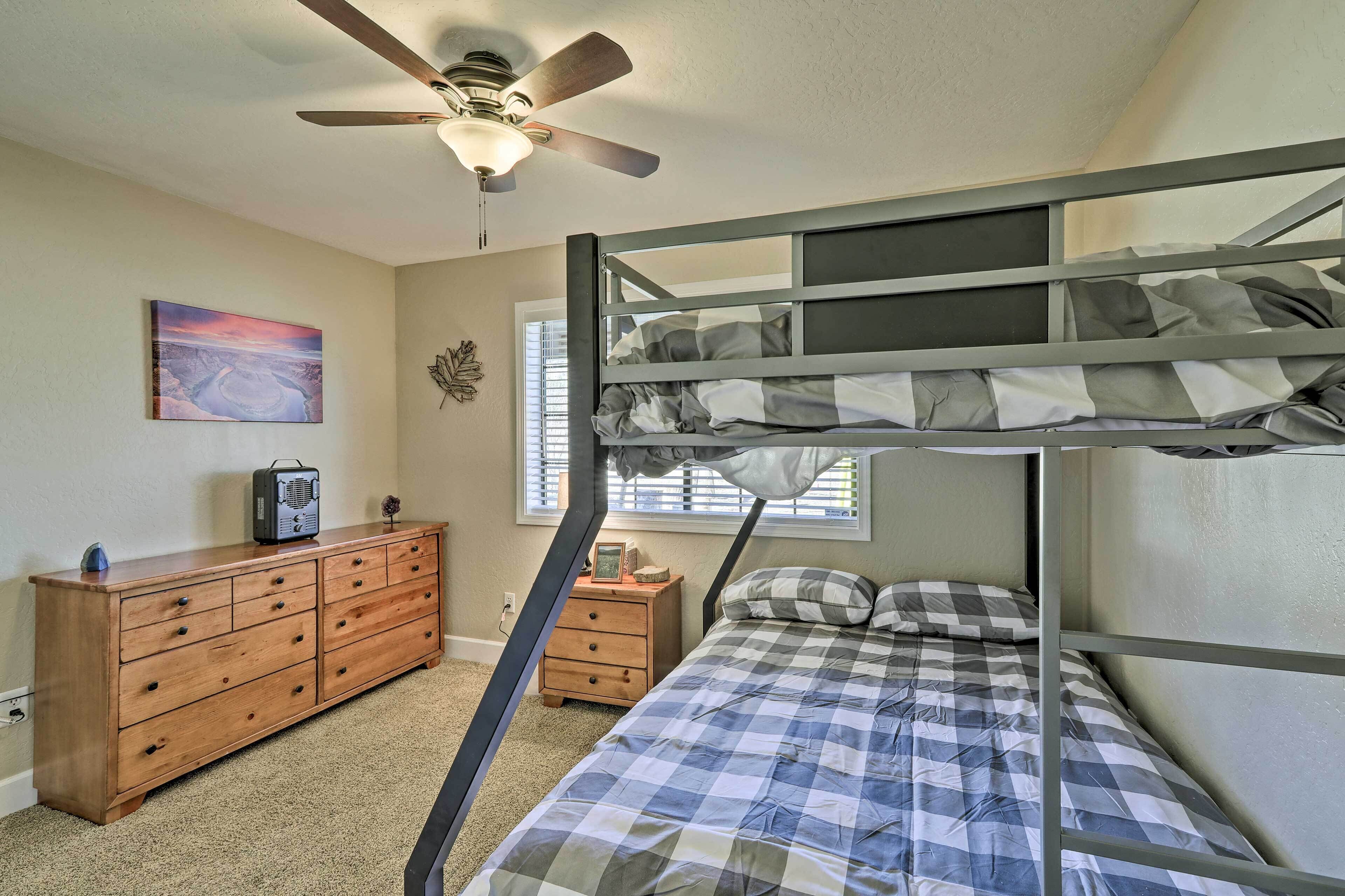 Une chambre avec un lit superposé, un ventilateur de plafond, une commode en bois, un tableau et une fenêtre avec des stores.