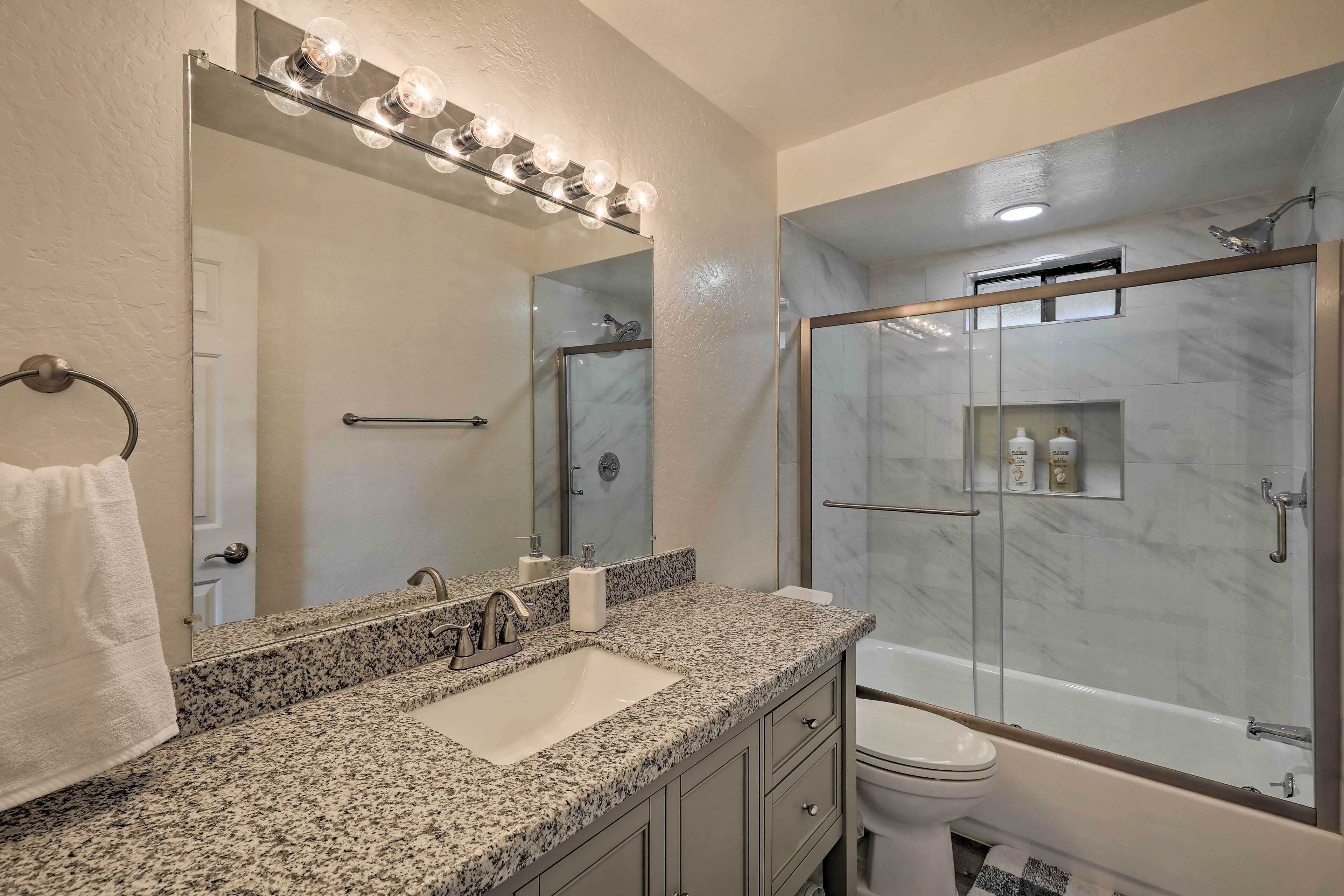 A bathroom with a granite countertop, a sink, a mirror with lights, a toilet, and a glass-enclosed shower area.