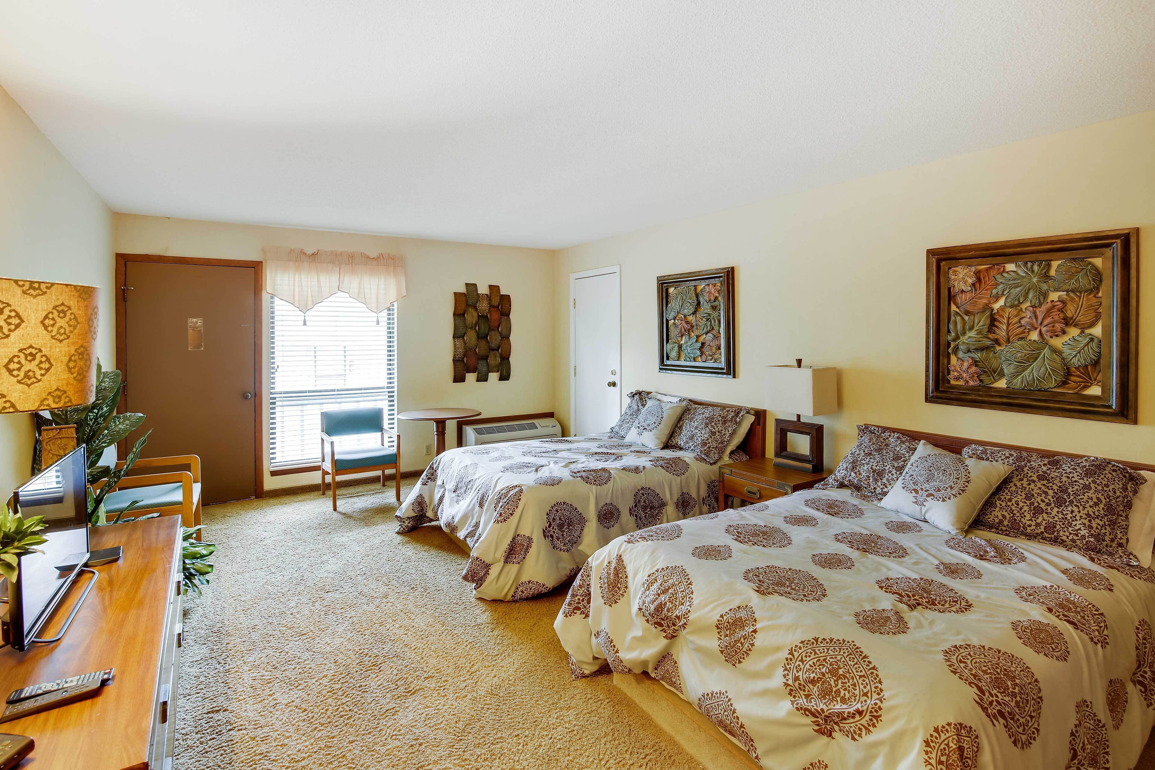 A hotel room with two beds, a desk, a chair, a small table, and a window with blinds.