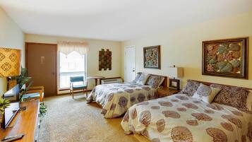 A hotel room with two beds, a desk, a chair, a small table, and a window with blinds.