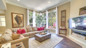 Un salon avec un canapé beige, un pouf en cuir, une table basse en bois, un téléviseur à écran plat et une grande fenêtre avec des rideaux.
