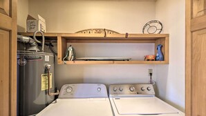 A laundry room with two white front-loading washers and dryers, a gas control valve, and a shelf above holding a flat iron, a spray bottle, and a box. - 'rocky Mountain Breezes' Cabin: Grill + Deck! (Estes Park)