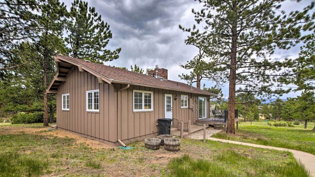 A small, single-story cabin with a brown exterior, white trim, and a chimney. It is surrounded by a grassy area with trees and a paved path leading to the entrance. - 'rocky Mountain Breezes' Cabin: Grill + Deck! (Estes Park)