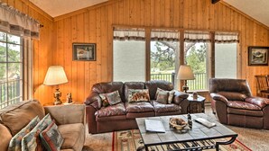 A cozy living room with a brown leather sofa, a brown armchair, and a coffee table. There are two windows with curtains, a lamp on a side table, and a framed picture on the wall. - 'rocky Mountain Breezes' Cabin: Grill + Deck! (Estes Park)