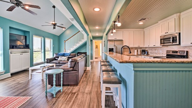 A modern kitchen with white cabinets, a stainless steel microwave, and a built-in oven. There is a bar area with stools and a seating area with a sofa and coffee table.