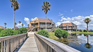 A wooden boardwalk leading to a building with a red roof, surrounded by palm trees and a calm body of water.