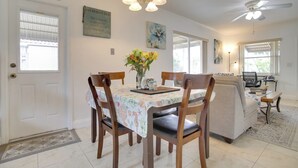 Un comedor con una mesa dispuesta para cuatro personas, rodeada de sillas de madera. Un jarrón con flores está sobre la mesa, y hay decoraciones en la pared, incluyendo una cita y un cuadro.