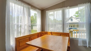 Ein Essbereich mit Holztisch und Bank, großen Fenstern mit leichten Vorhängen und Blick auf ein Haus und ein Auto draußen.