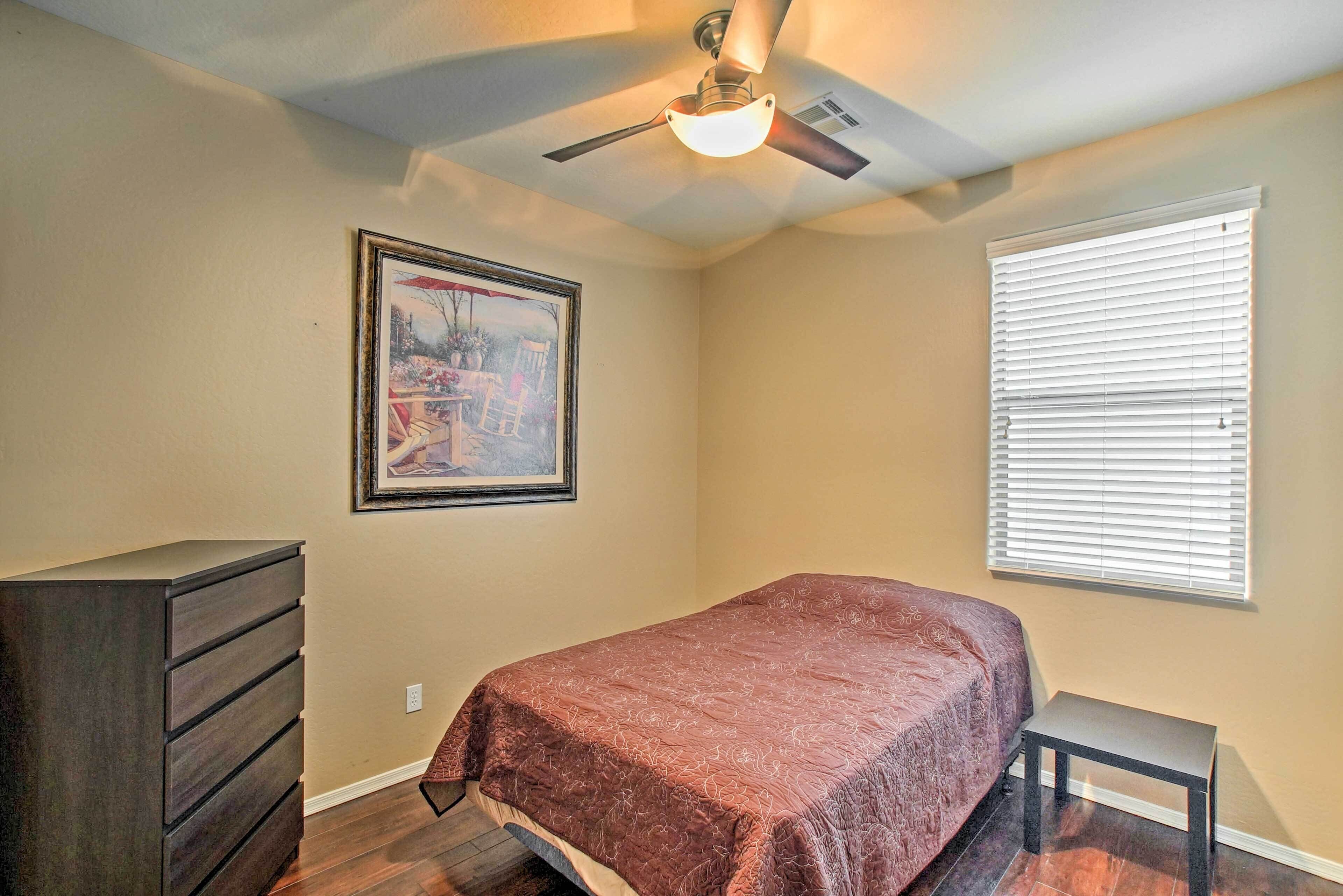 Une chambre à coucher comprenant un lit, une commode, un ventilateur de plafond, une fenêtre avec des stores et un tableau encadré accroché au mur.