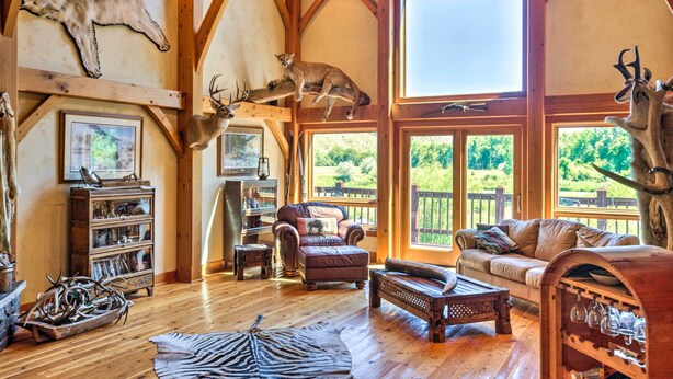 A spacious living room with wooden floors, a leather sofa, a wooden coffee table, and a large window offering a view of the outdoors. - Stunning Silesia Home: Furnished Deck + River View (Joliet)