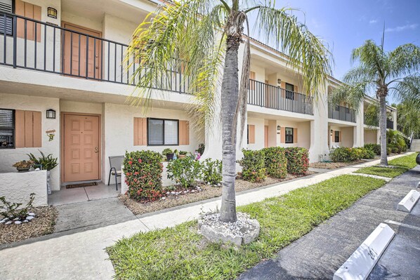 A multi-story residential building with balconies, a front door, and a small garden area with plants and a palm tree.