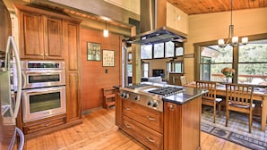 A kitchen with wooden cabinets, a built-in oven, a stove, and a dining area with wooden chairs and a table. - 9 Mi to Sugar Mountain: Stunning Cabin w/ Views (Banner Elk)