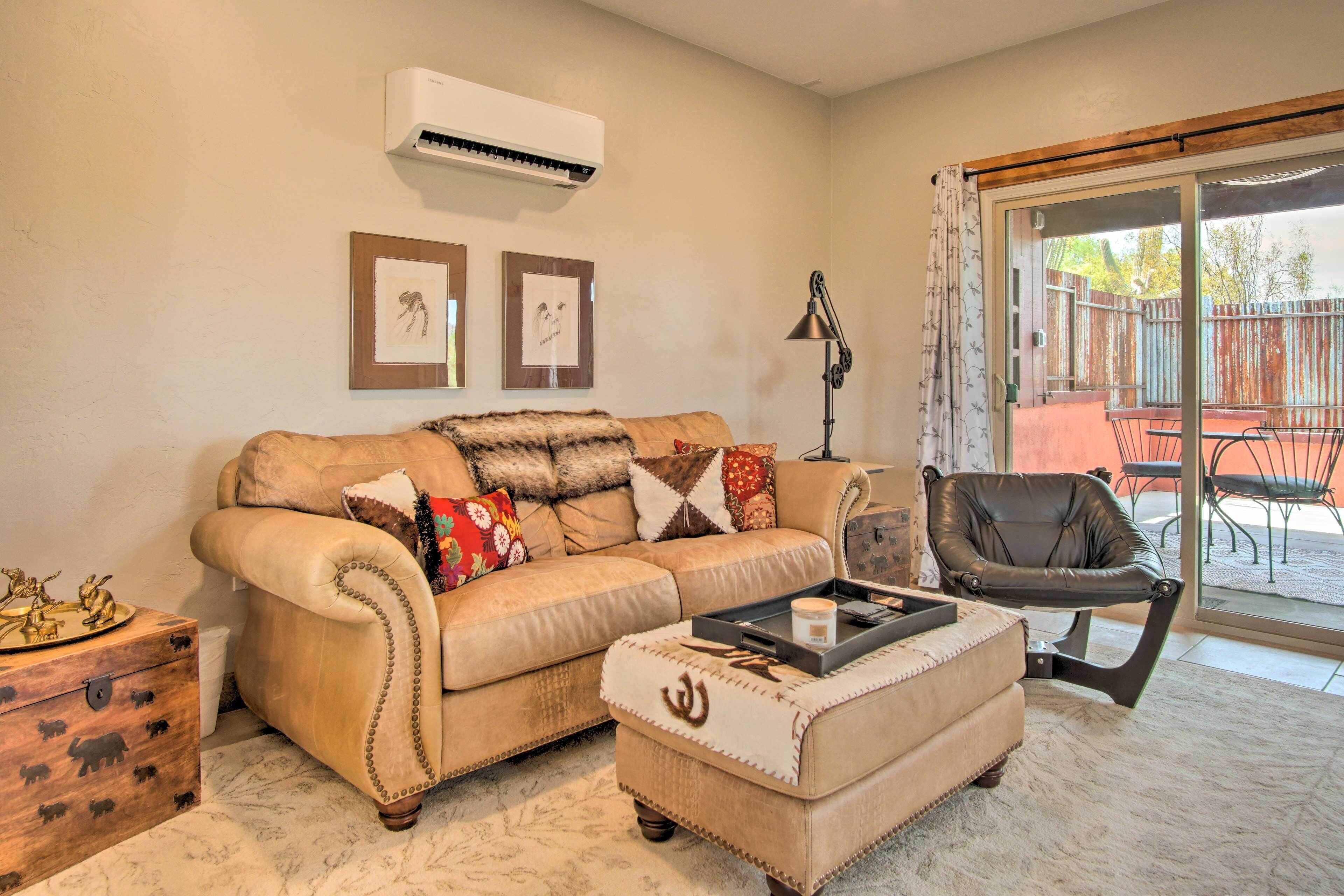 A living room with a beige sofa, a coffee table, and a leather chair. There is a sliding glass door leading to an outdoor patio.