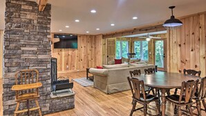 A cozy living room with a stone fireplace, wooden walls, a large window, and a dining area with a wooden table and chairs. - Rustic Sunset Escape on 67 Acres: Outdoor Kitchen (Sunset)