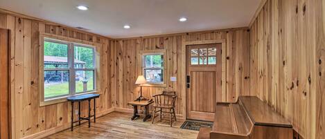 Une pièce aux murs lambrissés, avec un plancher en bois, un piano, une table avec une lampe et deux fenêtres donnant sur un paysage verdoyant.