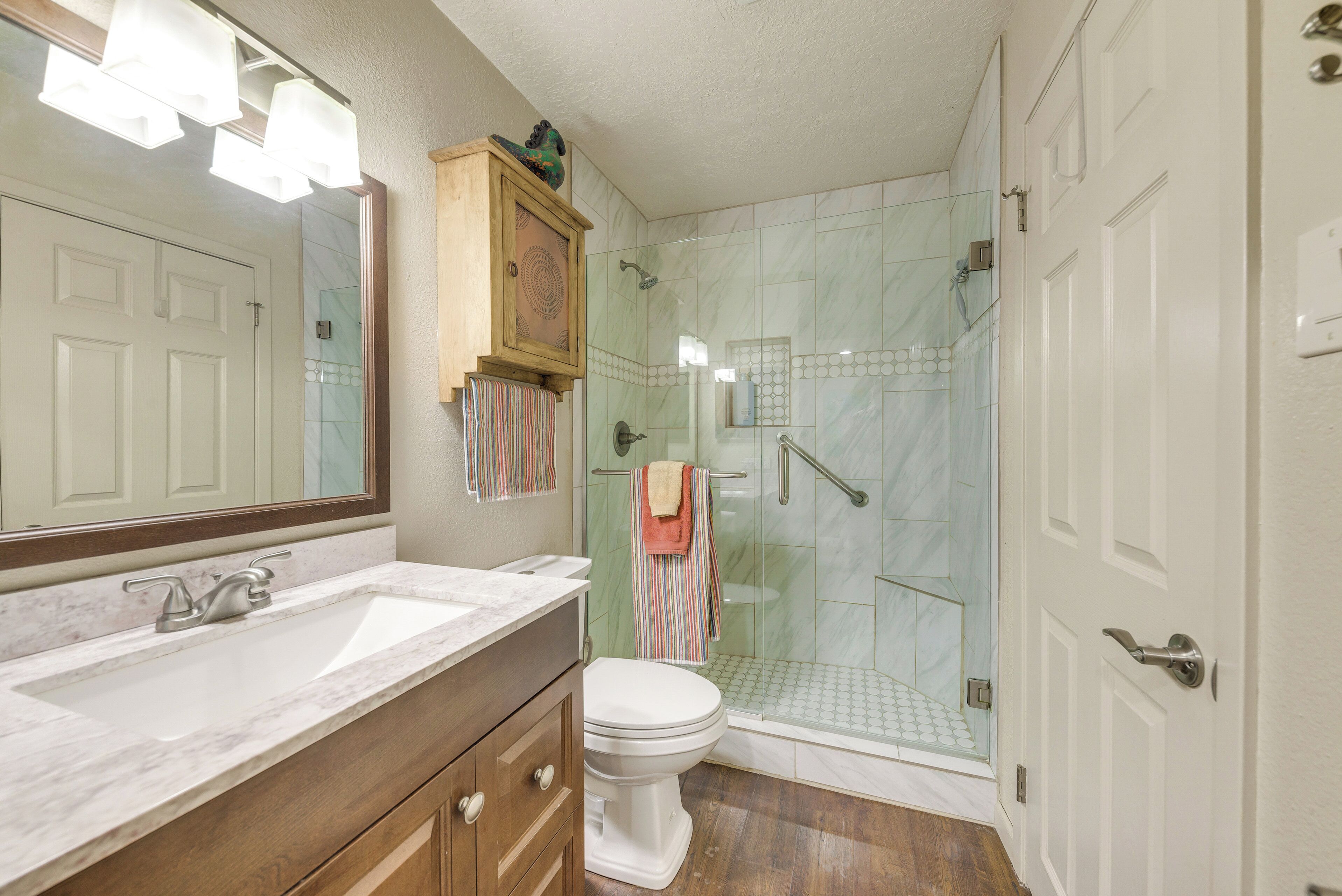 A bathroom with a marble sink, wooden vanity, a toilet, a glass shower enclosure, and a large mirror.