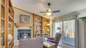A cozy living room with a fireplace, wooden shelves, a chessboard, and a view of the outdoors through large sliding doors.