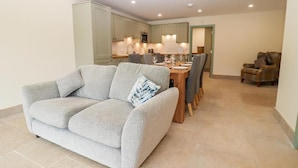 A living room with a grey sofa, a dining table with chairs, and a kitchen in the background. - 3 Bears Court Barn Conversion in Little Rissington (Cheltenham)