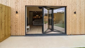 A modern building with a wooden exterior and a sliding glass door leading to an interior space with a sofa and a dining area. - 3 Bears Court Barn Conversion in Little Rissington (Cheltenham)
