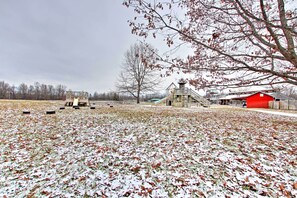 A playground with slides and climbing structures, a red barn, and a large tree in a snowy field.